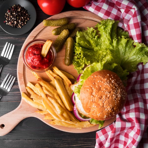 Top-down view of a burger, fries, ketchup, and pickles on a wooden board with a red checkered napkin.