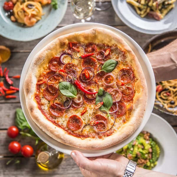 Hands serving a pepperoni pizza with red onions, chili peppers, and basil on a wooden table.