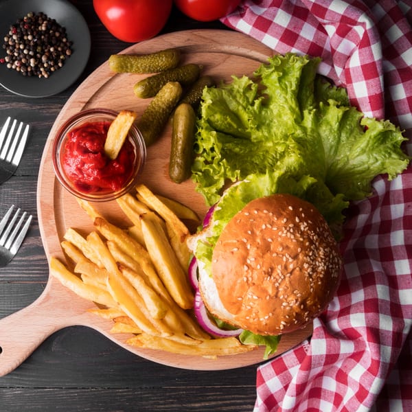A hamburger with fries, ketchup, pickles, and lettuce on a wooden board with a red checkered napkin.