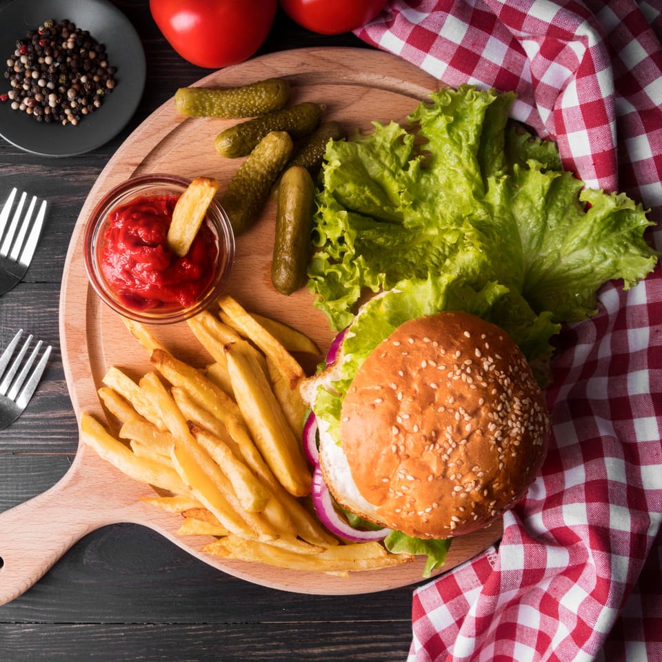 A hamburger with fries, ketchup, pickles, and lettuce on a wooden board with a red checkered napkin.