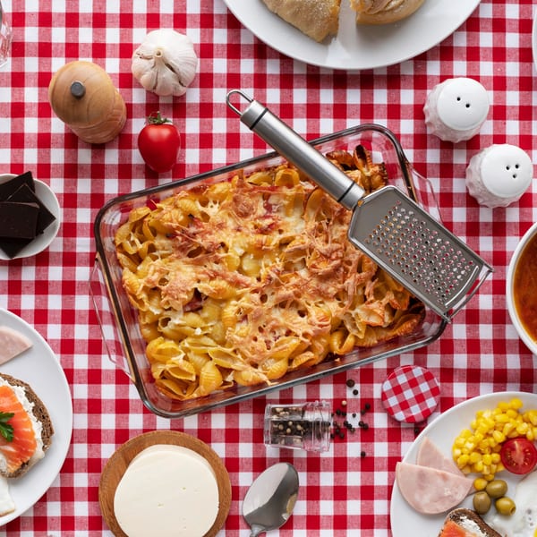 Baked pasta with melted cheese and a grater on a red and white checkered tablecloth.