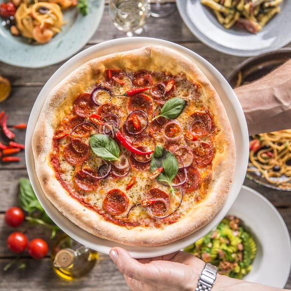 A hand holding a Roma's Pizza with pepperoni, red onion, chili, and basil, surrounded by other Italian dishes.