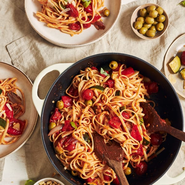 A large pot of Roma's Italian pasta with tomatoes, zucchini, olives, and basil, with plates nearby.